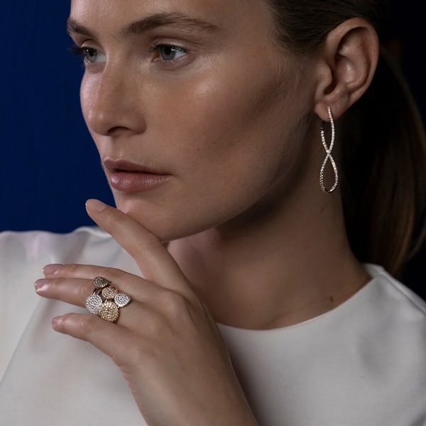 Eine Frau mit heller Haut und braunem Haar trägt ein weißes Oberteil und präsentiert eleganten Schmuck. Ihre Bigli-Ohrringe baumeln mit einem Anhänger in Unendlichkeitsform und mehrere Edelsteinringe schmücken ihre Finger. Vor einem dunkelblauen Hintergrund wirkt sie nachdenklich.