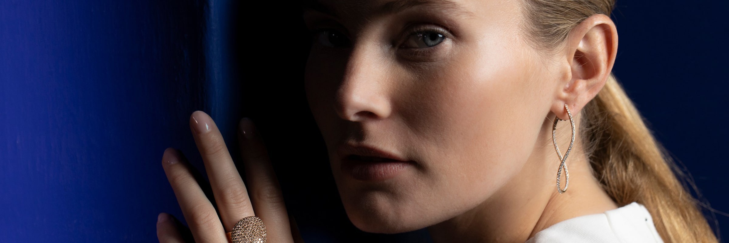 Eine Frau mit heller Haut und blondem Haar blickt zur Seite. Sie trägt ein Paar zarte Ohrringe und einen Ring mit strukturiertem Design. Ihre Hand liegt sanft auf einer dunkelblauen Oberfläche in sanftem Licht.