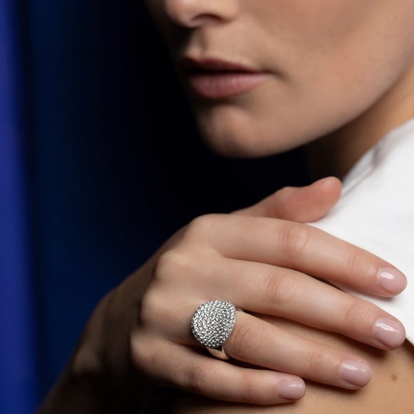 Eine Nahaufnahme der Hand einer Frau, die auf ihrer Schulter ruht, zeigt einen großen, funkelnden silbernen Bigli-Ring mit aufwendigen Details. Der Hintergrund ist eine Mischung aus dunkelblauen und weißen Tönen.