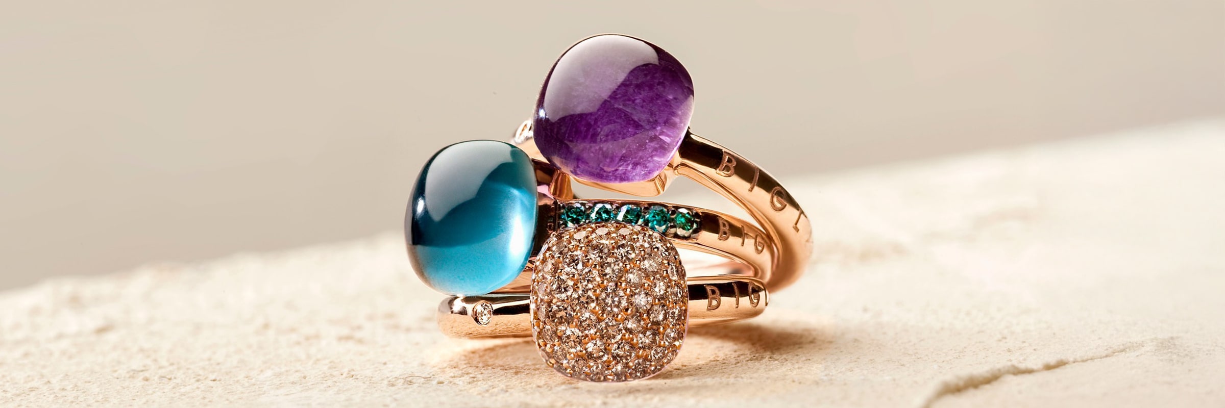 Drei elegante Ringe mit einzigartigem Design sind übereinander gestapelt. Der obere Ring hat einen glatten violetten Stein, der mittlere einen glänzenden blauen Stein und der untere zeigt eine funkelnde Oberfläche mit zahlreichen kleinen Edelsteinen. Die Bänder haben einen warmen, goldenen Farbton.