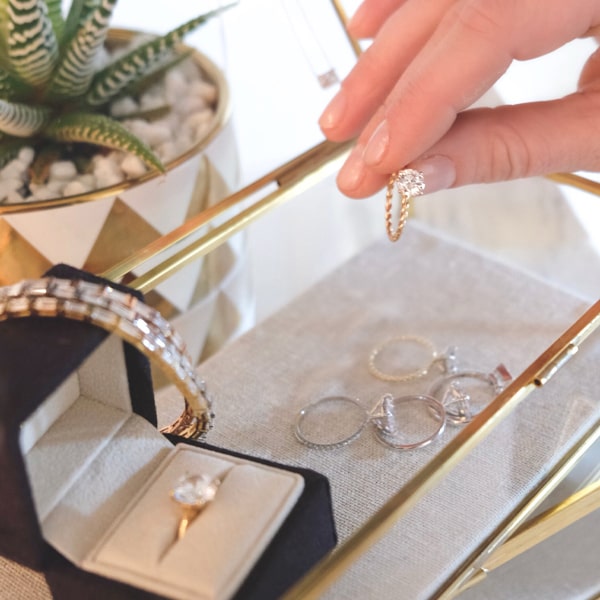 Diavon © 2024 Eine Person hält einen Diavon-Diamantring über einem Schmucktablett aus Glas, auf dem sich verschiedene Ringe und ein goldenes Armband befinden. Außerdem ist eine gepolsterte Ringschachtel mit einem Diamantring zu sehen. Im Hintergrund ist eine kleine grüne Sukkulente in einem gold-weißen Topf zu sehen.