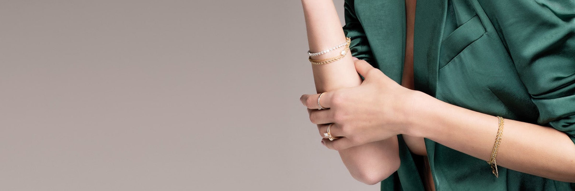 Nahaufnahme einer Person, die eine grüne Bluse mit hochgekrempelten Ärmeln trägt. An ihrem rechten Handgelenk sind mehrere goldene und silberne Armbänder zu sehen und an ihrer linken Hand ein goldener Ring. Der Hintergrund ist in neutralem Grau gehalten.