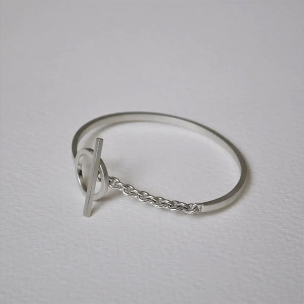 Ein minimalistisches Silberarmband von DOPPELLUDWIG Armschmuck, bestehend aus einem dünnen Metallband, das mit einem kleinen runden Ring und einem horizontalen Balken verbunden ist. Das Armband enthält ein kurzes Kettenstück, das Band und Ring verbindet. Elegant präsentiert auf einer schlichten weißen Oberfläche.