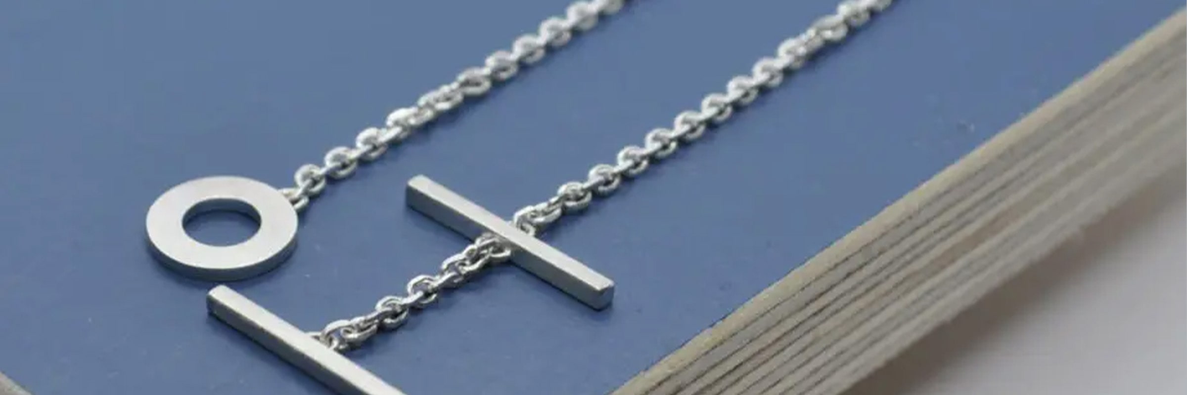 Eine Nahaufnahme einer Silberkette auf einer blauen Oberfläche. Die Kette hat zwei geometrische Anhänger, einen runden und einen vertikalen Balken. Die Gegenüberstellung der Anhänger ergibt ein minimalistisches und modernes Design.