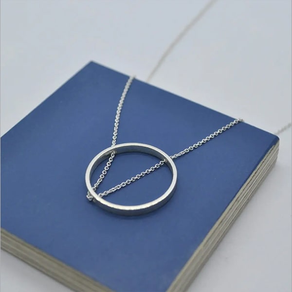 Eine zarte DOPPELLUDWIG-Silberkette mit rundem Anhänger wird elegant auf einer blauen quadratischen Box präsentiert. Das minimalistische Anhängerdesign verfügt über ein dünnes, glattes rundes Band, durch das die Kette der Kette verläuft. Die Silberkette trägt zum schlichten und doch raffinierten Look bei.