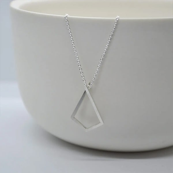Doppelludwig © 2024 Eine minimalistische, silberne Halskette von DOPPELLUDWIG Halsschmuck mit einem leeren, rautenförmigen Anhänger ist elegant über den Rand einer glatten, weißen Keramikschale drapiert. Der Anhänger zeichnet sich durch klare Linien und ein schlichtes Design aus und erzeugt vor dem schlichten Hintergrund ein modernes und stilvolles Erscheinungsbild.