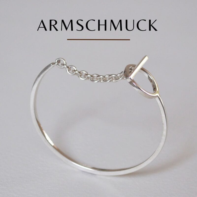 Vor einem schlichten Hintergrund befindet sich ein minimalistisches Silberarmband mit Kette und rundem Verschluss. Darüber steht in fetten Lettern das Wort „ARMSCHMUCK“.
