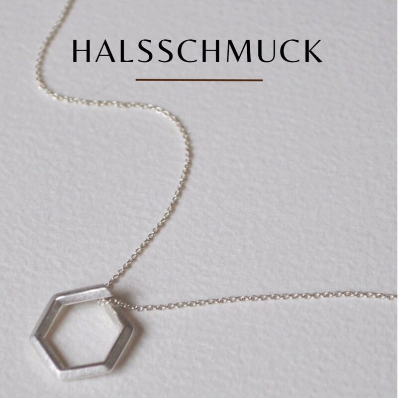 Auf hellem Untergrund liegt eine zarte Silberkette mit sechseckigem Anhänger. Darüber steht in Großbuchstaben das Wort „HALSSCHMUCK“.