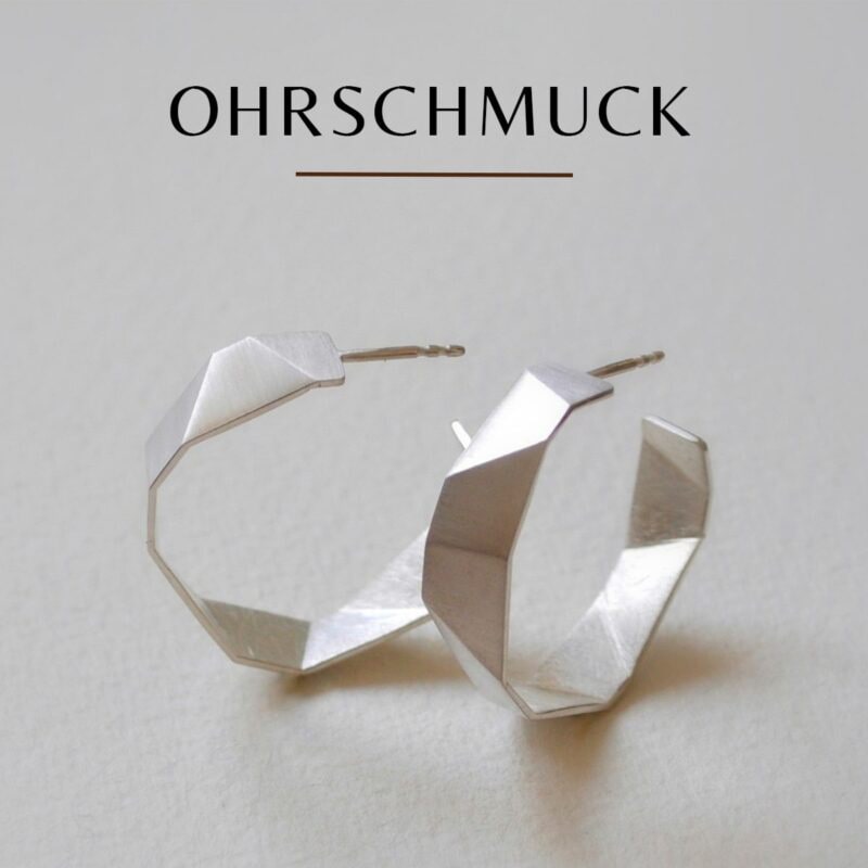 Ein Paar geometrische, Origami-inspirierte Creolen aus Silber auf glatter Oberfläche. Über den Ohrringen steht in fetten schwarzen Buchstaben das Wort „OHRSCHMUCK“.