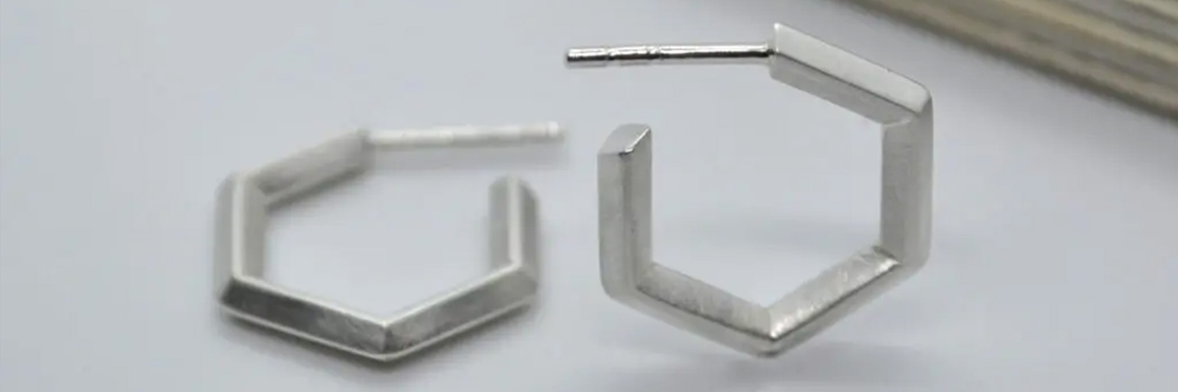 Ein Paar silberne sechseckige Ohrringe wird ausgestellt. Ein Ohrring steht aufrecht und der andere liegt flach. Die Ohrringe haben ein schlichtes und minimalistisches Design mit einer modernen geometrischen Form. Sie sind vor einem einfachen, hellen Hintergrund angebracht.