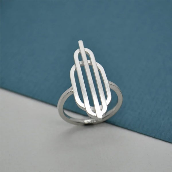Ein minimalistischer Silberring mit abstraktem geometrischem Design, bestehend aus parallelen Linien und überlappenden Formen von DOPPELLUDWIG. Der Ring ist vor einem geteilten Hintergrund in Blau und Weiß fotografiert.