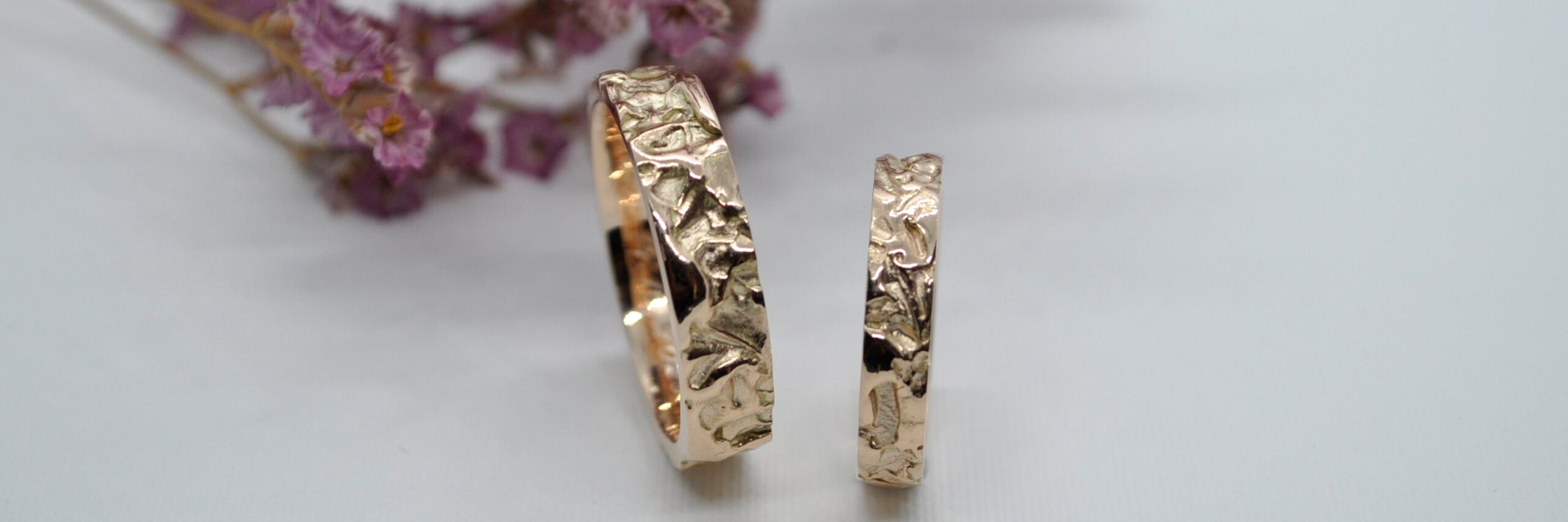 Zwei aufwendig gestaltete Goldringe mit strukturiertem Muster liegen nebeneinander auf einer weißen Oberfläche. Der größere Ring ist links und der kleinere rechts. Im Hintergrund sind unscharf Zweige hellvioletter Trockenblumen zu sehen.
