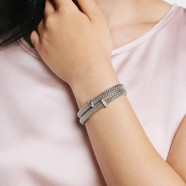 FOPE © 2024 Eine Person, die eine hellrosa Bluse trägt, ist von den Schultern abwärts zu sehen und richtet mit der rechten Hand ein silbernes geflochtenes Fope-Armband. Der Hautton der Person ist hell und ihre Nägel sind natürlich und nicht lackiert. Der Hintergrund ist schlicht und größtenteils weiß.