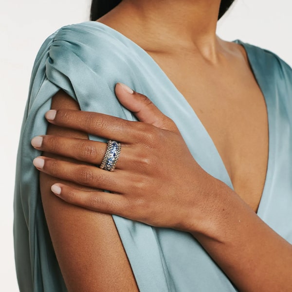 FOPE © 2024 Eine Person in einem hellblauen Satinkleid zeigt ihre Hand, die mit einem breiten, mehrfarbigen Edelsteinring geschmückt ist. Die andere Hand der Person berührt sanft den Arm, um den Schmuck hervorzuheben. Dies zeigt einen eleganten und stilvollen Look, der leicht mit einem Stück aus Fopes Kollektion akzentuiert werden könnte.
