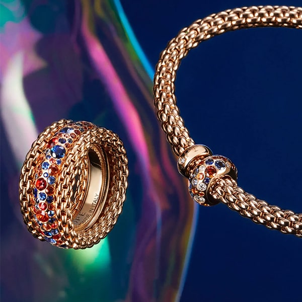 FOPE © 2024 Eine Nahaufnahme eines verzierten Fope-Goldrings und eines passenden Armbands vor einem farbenfrohen, schillernden Hintergrund. Beide Stücke weisen komplizierte gewebte Muster auf und sind mit kleinen roten, blauen und klaren Edelsteinen verziert, die in einem eleganten Design angeordnet sind.