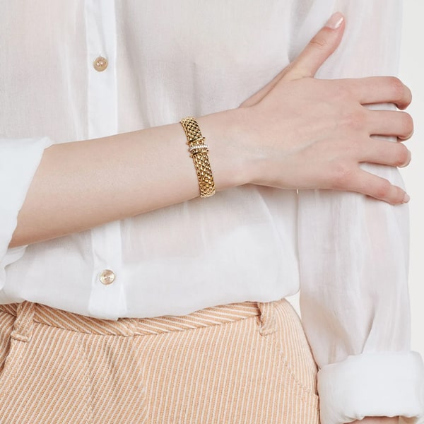 FOPE © 2024 Eine Person in einem weißen Langarmhemd und einer beigen Nadelstreifenhose präsentiert ein goldenes Kettenarmband von Fope Armschmuck mit einem kleinen Schnallendetail. Das Armband wird am linken Handgelenk getragen, während die rechte Hand sanft auf dem Unterarm ruht.