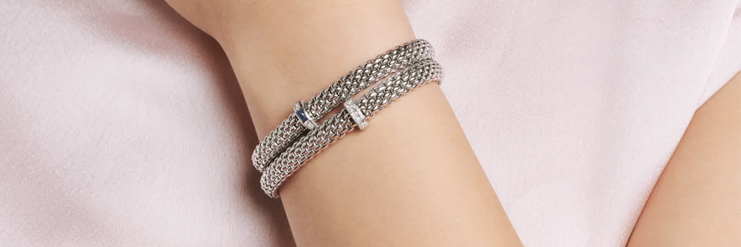 Eine Nahaufnahme des Arms einer Person, die ein mehrreihiges Silberarmband trägt, das mit kleinen, zarten Anhängern verziert ist. Das Armband ist mehrmals um das Handgelenk gewickelt, wodurch ein mehrschichtiger Effekt entsteht. Die Person trägt ein hellrosa Kleidungsstück.