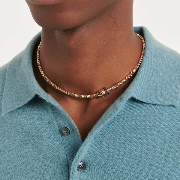 Nahaufnahme einer Person, die Fope Halsschmuck trägt, eine strukturierte goldene Halskette mit Emaille-Detail. Die Person trägt ein hellblaues Hemd mit Kragen. Die Teilansicht zeigt nur die untere Hälfte ihres Gesichts und den Oberkörper.