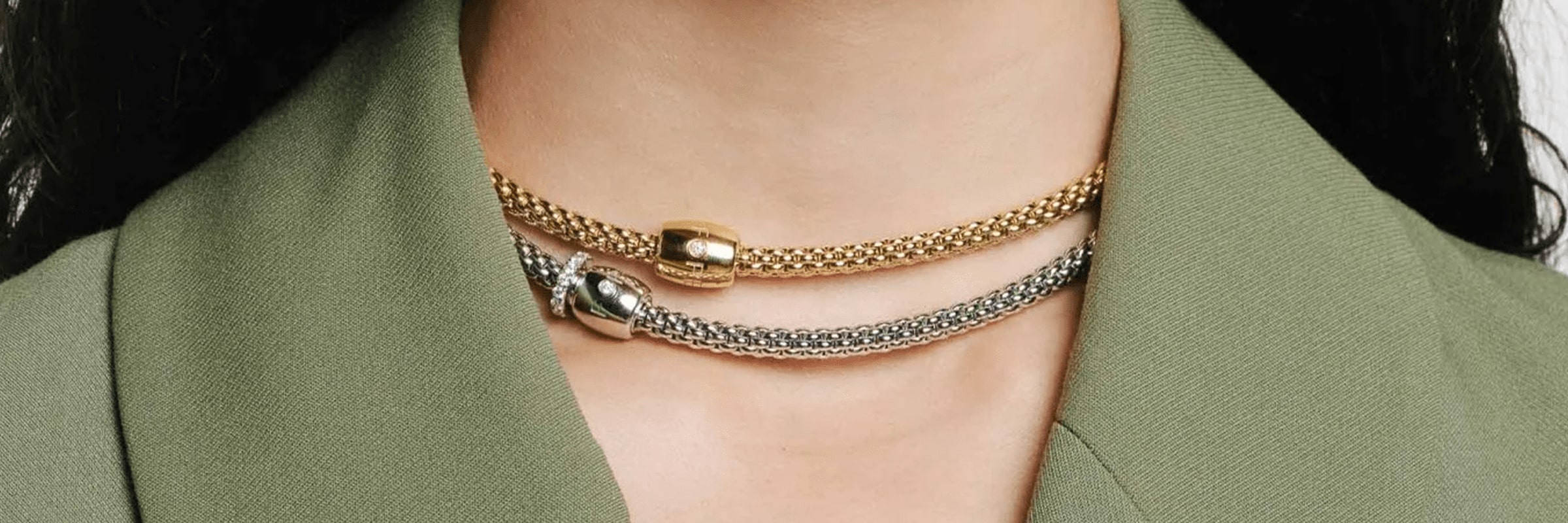 Nahaufnahme des Halses einer Person, die einen grünen Blazer trägt und mit zwei grob gewebten Halsbändern geschmückt ist. Das obere Halsband ist golden mit einer zentralen Metallperle und das untere Halsband ist silberfarben mit einer glänzenden runden Perle.