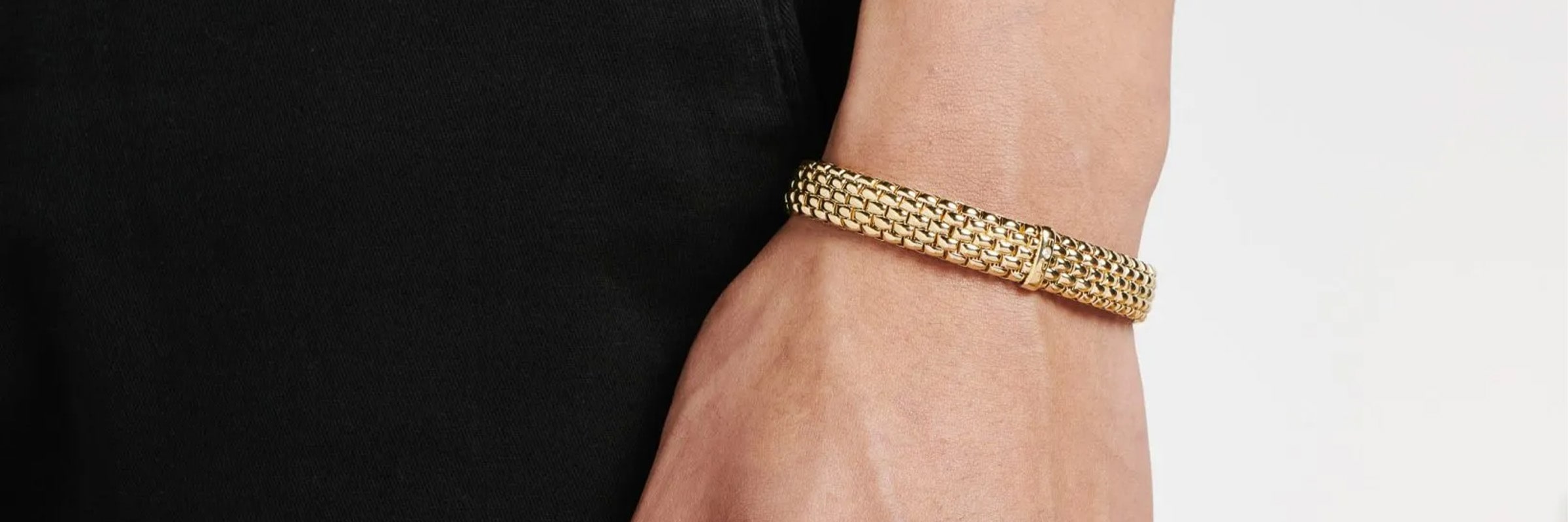Eine Nahaufnahme des Handgelenks einer Person, die mit einem strukturierten goldenen Armband geschmückt ist. Die Person trägt ein schwarzes Kleidungsstück, das einen Kontrast zum goldenen Armband bildet. Das Armband hat ein kompliziertes, gewebtes Design.
