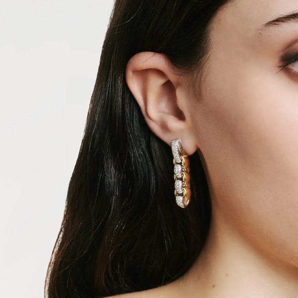 FOPE © 2024 Eine Nahaufnahme des Ohrs einer Person, die mit einem Fope Ohrschmuck-Creolen aus Gold und Diamanten geschmückt ist. Der Ohrring weist ein abwechselndes Muster aus Gold- und Diamantnieten auf. Die Person hat langes, dunkles Haar, das teilweise vor einem schlichten weißen Hintergrund sichtbar ist.