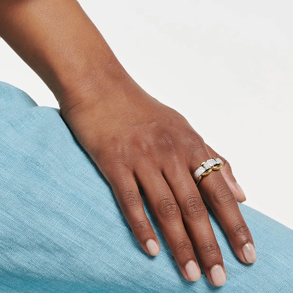 FOPE © 2024 Eine Nahaufnahme einer Hand, die auf einer hellblauen Stoffoberfläche ruht. Die Hand ist mit einem Fope-Ring geschmückt, in dessen Mitte sich ein großer, funkelnder Edelstein befindet, der von zwei kleineren Edelsteinen flankiert wird. Die Nägel sind kurz gehalten und sorgfältig manikürt.