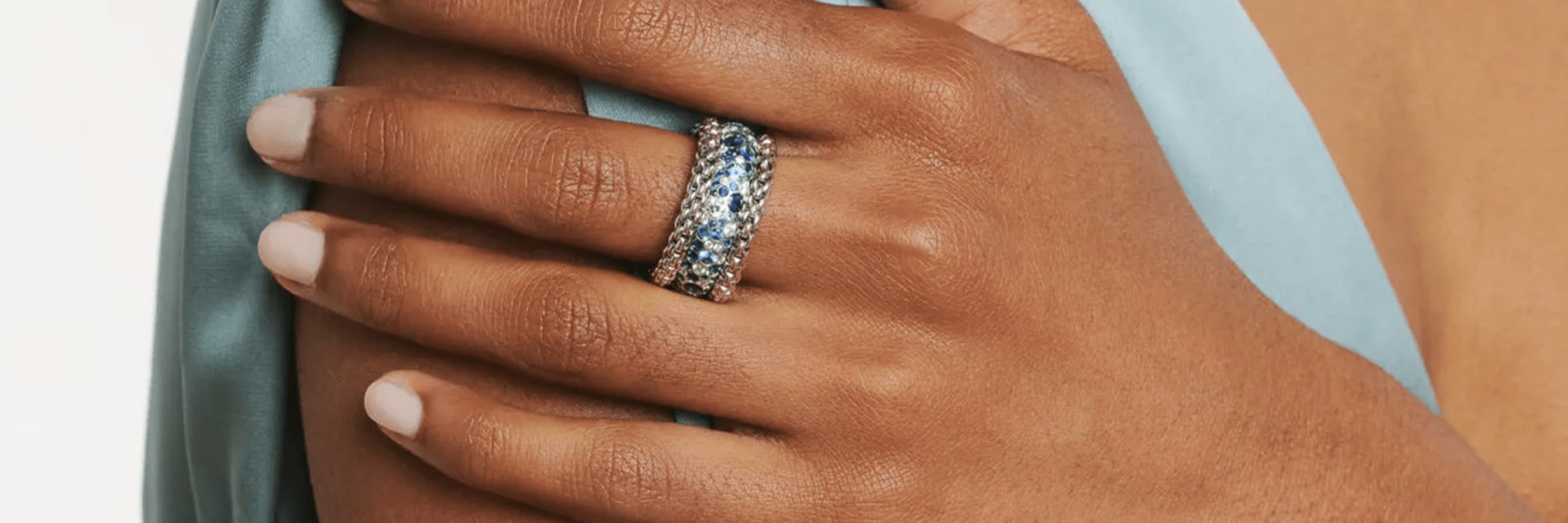 Eine Hand mit hellrosa Nagellack ruht auf einem hellblauen Stoff. Die Hand ist mit einem dicken, mehrfarbigen Edelsteinring geschmückt. Der Stoff ist um die Schulter drapiert und suggeriert eine elegante oder formelle Umgebung.