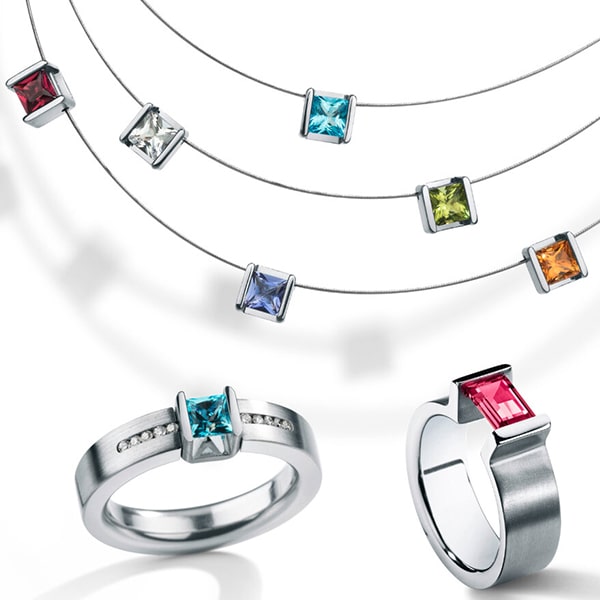 Humphrey © 2024 Eine Kollektion von Humphrey-Silberschmuck mit quadratisch geschliffenen Edelsteinen. Drei Halsketten mit verschiedenfarbigen Steinen, darunter rote, klare, blaue, grüne und orange, sind über zwei Ringen zu sehen. Ein Ring hat einen blauen Edelstein mit kleinen Diamanten auf dem Band, während der andere einen roten Edelstein zur Schau stellt.