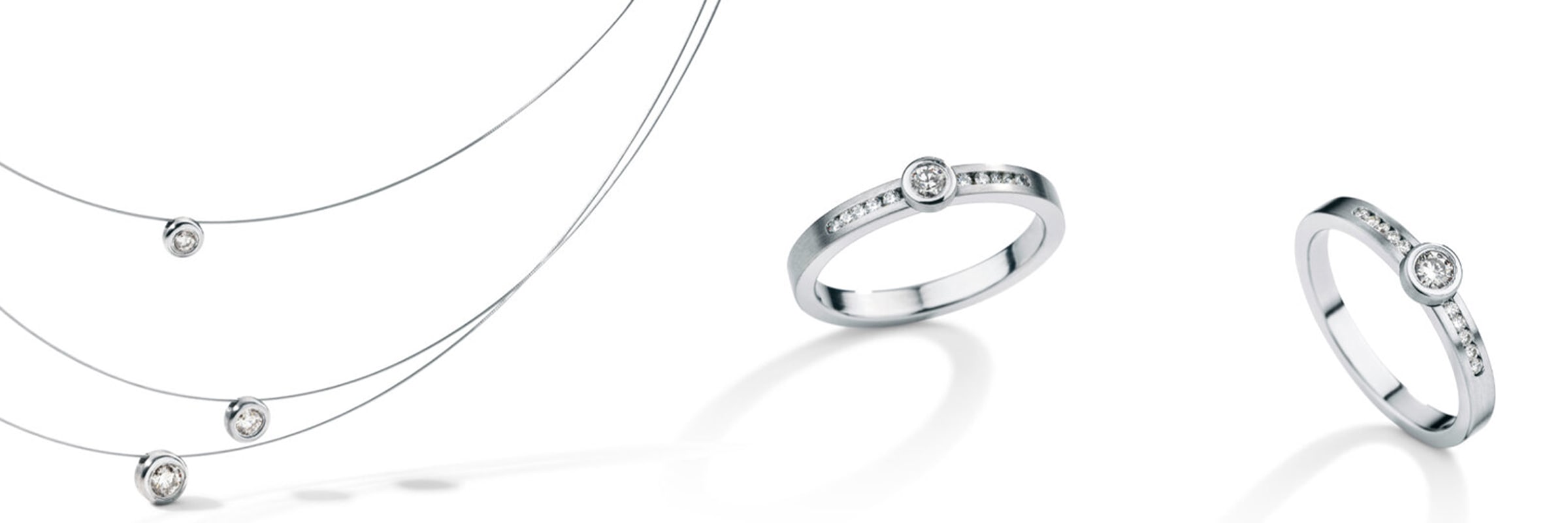 Drei Silberketten mit jeweils einem einzelnen Diamantanhänger, der in unterschiedlicher Länge herabhängt, und drei Silberringe mit eingelegten Diamanten, die ordentlich daneben auf einem weißen Hintergrund angeordnet sind.