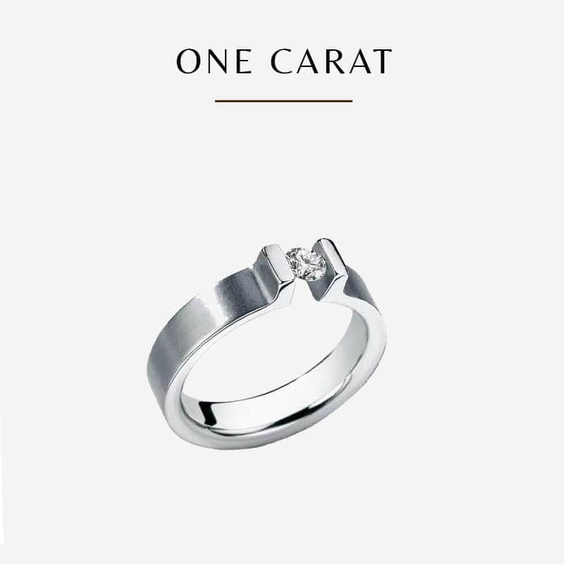Eine Nahaufnahme eines silbernen Humphrey-Verlobungsrings mit einem einzelnen Diamanten vor einem schlichten weißen Hintergrund. Über dem Ring steht in eleganten Großbuchstaben „EIN KARAT“.