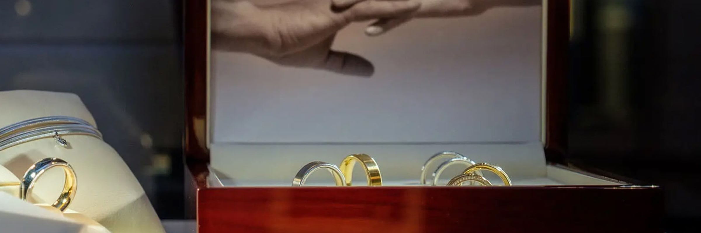 In einem Juweliergeschäft wird eine Auswahl an Gold- und Silberringen präsentiert. Eine Samtschachtel mit mehreren Ringen ist geöffnet, im Hintergrund ist ein Schwarzweißfoto eines Händchen haltenden Paares zu sehen. Weitere Ringe in einer kleineren Vitrine sind links teilweise sichtbar.