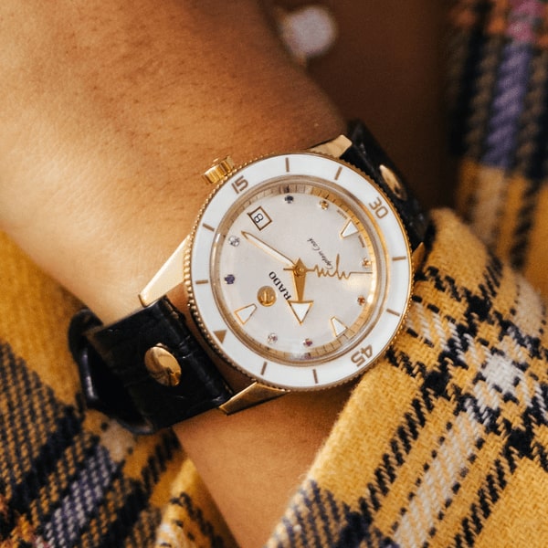 Nahaufnahme eines Handgelenks, das mit einer eleganten gold-weißen analogen Armbanduhr von Uhren geschmückt ist, ergänzt durch ein schwarzes Lederarmband. Das Zifferblatt verfügt über goldene Stundenmarkierungen und Zeiger, ein Datumsfenster und dekorative Elemente. Das Handgelenk ist in einen gemusterten gelb-schwarzen Stoff gehüllt.