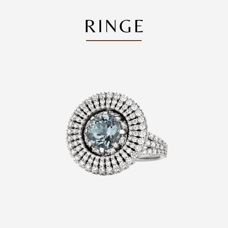 Ein luxuriöser Heinz-Ring mit einem zentralen blauen Edelstein, der von mehreren Reihen kleiner weißer Diamanten umgeben ist. Das Band ist ebenfalls mit Diamanten verziert. Über dem Ring ist das Wort „Ringe“ in einer stilvollen Schriftart abgebildet. Der Hintergrund ist in minimalistischem Weiß gehalten.