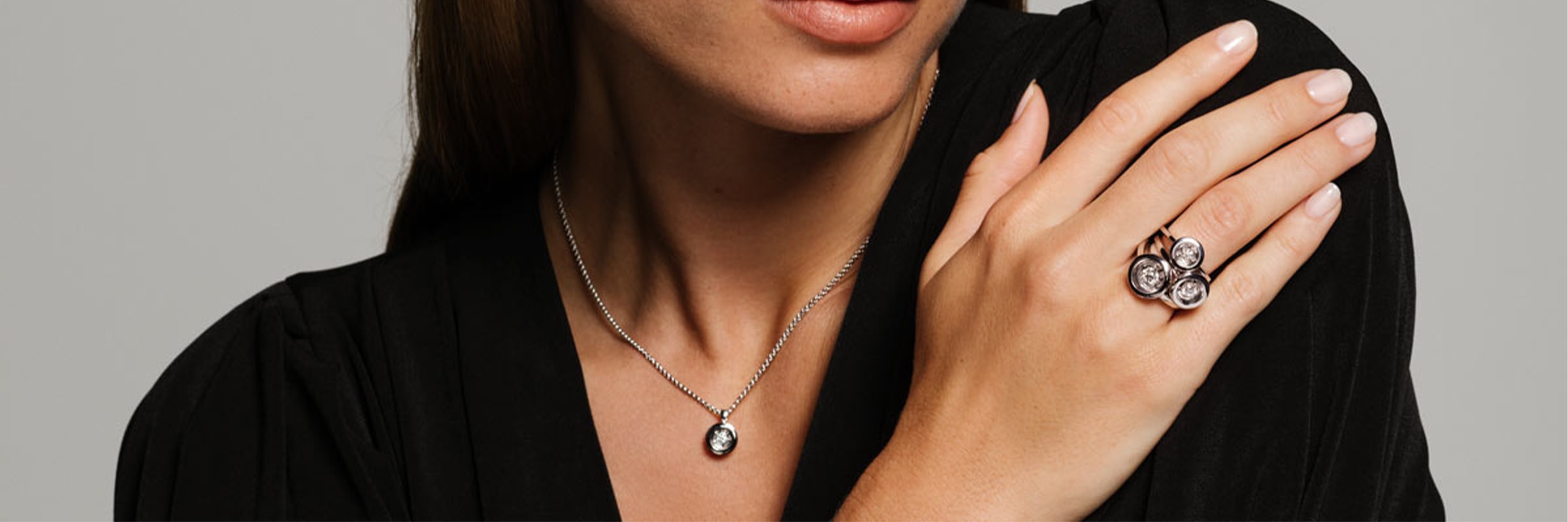 Eine Person trägt ein schwarzes Oberteil und trägt Accessoires wie eine Halskette mit einem runden Anhänger und einen Statement-Ring mit drei großen Steinen an ihrer rechten Hand. Der Kopf der Person ist teilweise sichtbar und leicht zur Seite gedreht.