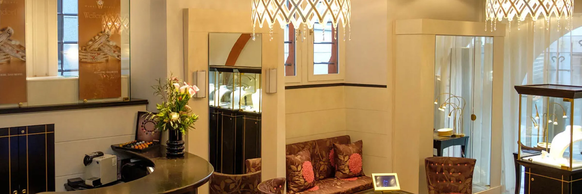 Ein luxuriöses Juweliergeschäft mit eleganten Kronleuchtern, Glasvitrinen mit Schmuck und einem gemütlichen Sitzbereich mit weichen Kissen und Blumen. Die Einrichtung umfasst sanfte Beleuchtung, geschmackvolle Wandkunst und eine Kaffeemaschine auf einer eleganten Theke.