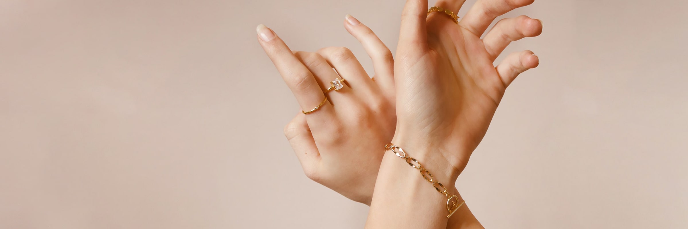 Zwei Hände, die anmutig mit leicht gespreizten Fingern posierten. Die linke Hand trägt einen goldenen Ring mit einem Edelstein, während die rechte Hand einen dünnen goldenen Ring trägt. Beide Handgelenke sind mit zarten goldenen Armbändern geschmückt. Der Hintergrund ist in sanftem Beige gehalten und schafft eine minimalistische und elegante Kulisse.
