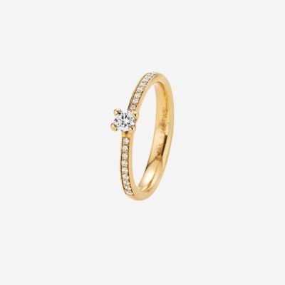 © Juwelier Wagner-Madler Ein Goldring mit einem einzelnen runden Diamanten in der Mitte und kleineren Diamanten entlang der Hälfte des Rings, die an Einträge in jedem Schmucklexikon erinnern. Der Ring wird aufrecht auf einem weißen Hintergrund mit sichtbaren Gravuren auf der Innenseite des Rings präsentiert.