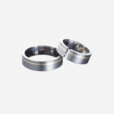 © Juwelier Wagner-Madler Ein Paar passender Silberringe, die jeweils einen dünnen Goldstreifen in der Mitte aufweisen. Ein Ring ist mit einem einzelnen kleinen Diamanten besetzt und beide Ringe haben eine glatte, polierte Oberfläche. Auf einem weißen Hintergrund positioniert, könnten diese exquisiten Stücke problemlos in jedes Schmucklexikon aufgenommen werden.