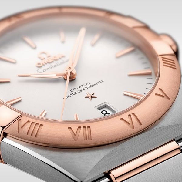 Nahaufnahme einer luxuriösen Omega Constellation-Armbanduhr. Die Uhr hat ein Gehäuse aus gebürstetem Edelstahl mit einer polierten Roségold-Lünette mit eingravierten römischen Ziffern. Das Zifferblatt ist weiß mit goldfarbenen Zeigern und Stundenmarkierungen und bei 6 Uhr befindet sich ein Datumsfenster.