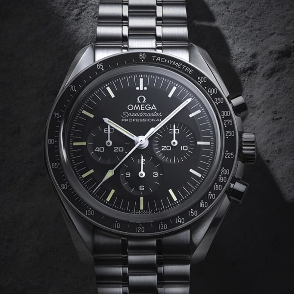 OMEGA © 2025 Eine klassische Omega Speedmaster Chronographenuhr mit Edelstahlgehäuse und -armband ist auf einer strukturierten, felsigen Oberfläche platziert. Die Omega-Uhr verfügt über ein schwarzes Zifferblatt mit drei Hilfszifferblättern und weißen Stundenmarkierungen.