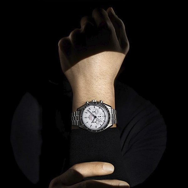 OMEGA © 2025 Eine silberne Armbanduhr aus der Omega Moonwatch Kollektion mit weißem Zifferblatt und mehreren Hilfszifferblättern wird am Handgelenk einer Person präsentiert. Die Uhr mit ihrem Metallarmband glänzt vor dem schwarzen Hintergrund, während die andere Hand der Person das Handgelenk zurechtrückt oder hält, und unterstreicht so ihre zeitlose Eleganz.