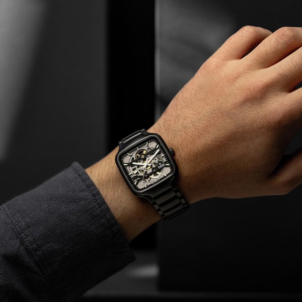 Ein Zeiger trägt eine schwarze Metallarmbanduhr mit sichtbarem mechanischem Innenleben, Zahnrädern und filigranen Details. Der dunkle und unscharfe Hintergrund unterstreicht das Design und die Handwerkskunst der Uhr.