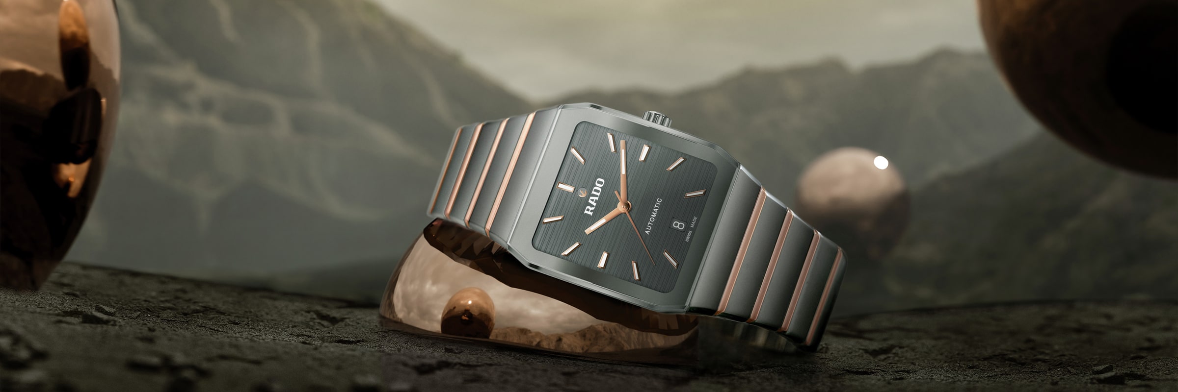 Eine elegante Armbanduhr mit Metallarmband präsentiert sich vor einer Bergkulisse mit reflektierenden Metallkugeln. Das Zifferblatt ist dunkel gehalten und weist schlichte Indizes sowie den Markennamen am oberen Rand auf.