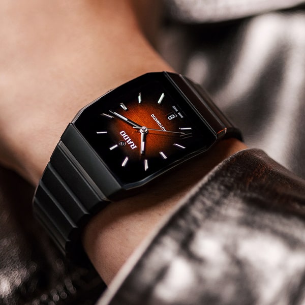 RADO © 2025 Eine Nahaufnahme eines Handgelenks, an dem eine elegante schwarze Metalluhr aus der Rado Anatom Kollektion getragen wird. Das Zifferblatt ist dunkel und weist einen Farbverlauf auf, der zur Mitte hin von Schwarz nach Orange übergeht. Das Datum wird auf der 6-Uhr-Position angezeigt und der Markenname „RADO“ unterhalb der 12-Uhr-Markierung.