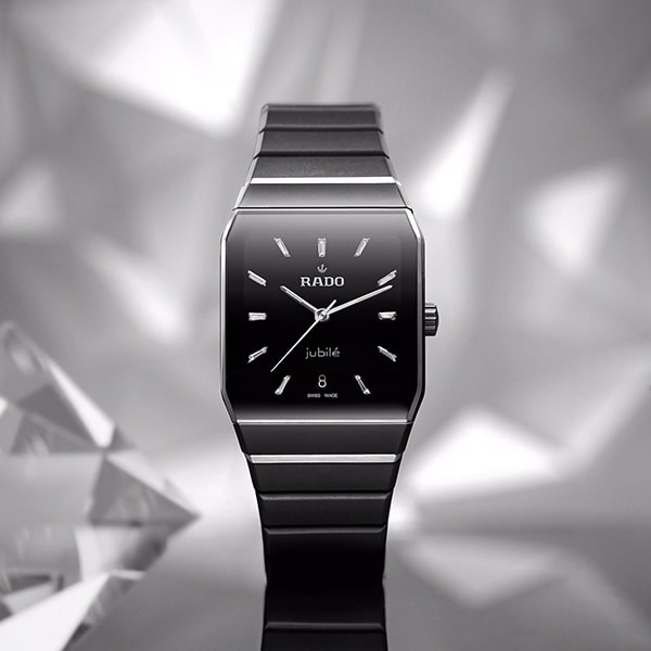 RADO © 2025 Eine Rado-Armbanduhr aus der Anatom Kollektion mit rechteckigem schwarzem Zifferblatt und silbernem Armband wird vor einem geometrischen, reflektierenden Hintergrund präsentiert. Die Uhr verfügt über silberne Stundenmarkierungen, schlanke Zeiger und ein Datumsfenster bei 6 Uhr. Auf dem Zifferblatt ist das Wort „Jubilé“ zu sehen.