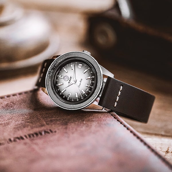 RADO © 2025 Eine stilvolle Armbanduhr aus der Rado Captain Cook Kollektion mit dunklem Lederarmband und rundem Zifferblatt aus Metall mit weißen Markierungen und Zeigern liegt auf einer Oberfläche aus braunem Leder. Der Hintergrund ist verschwommen und lenkt die Aufmerksamkeit auf die Uhr.