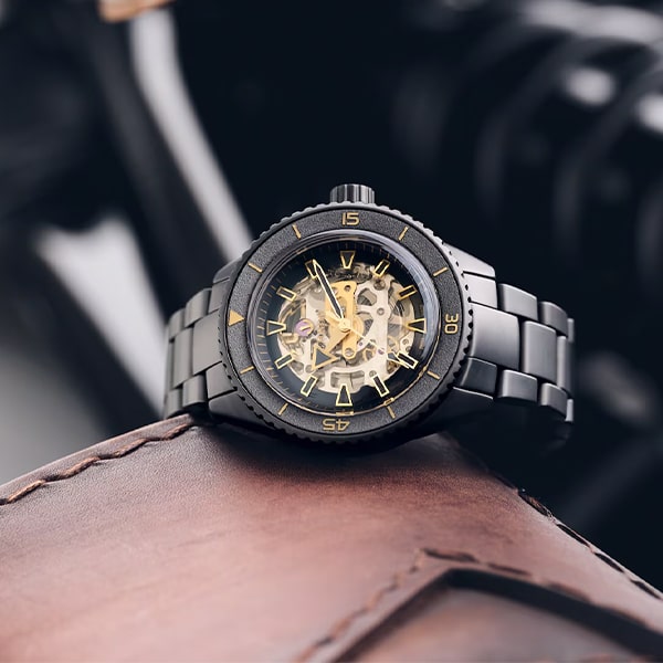 RADO © 2025 Eine schwarz-goldene Skelett-Armbanduhr aus der Rado Captain Cook Kollektion mit Metallarmband wird auf einer Lederoberfläche präsentiert. Die Uhr zeigt komplizierte innere Mechanismen und hat ein modernes, elegantes Design. Der Hintergrund ist verschwommen und hebt die Uhr als Mittelpunkt hervor.