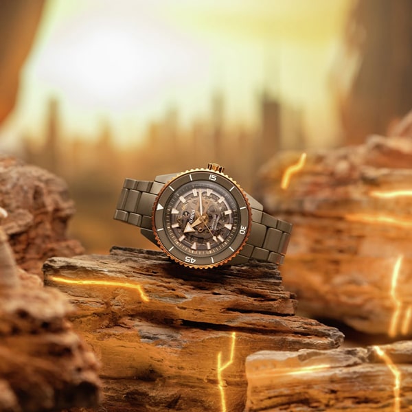 Eine robuste Armbanduhr mit dunklem Metallarmband ist vor einer wüstenähnlichen Kulisse zu sehen. Im Hintergrund sind leuchtende Felsformationen und eine verschwommene Skyline unter einem hellen, dunstigen Himmel zu sehen.