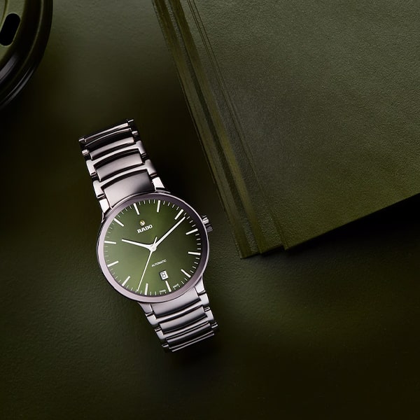 RADO © 2025 Eine elegante silberne Armbanduhr aus der Rado Centrix Kollektion mit grünem Zifferblatt und Metallarmband liegt auf einer grünen Oberfläche neben grün gefärbten Blättern oder Büchern. Die Uhr trägt den Markennamen „Rado“ auf dem Zifferblatt und weist ein minimalistisches und elegantes Design auf.