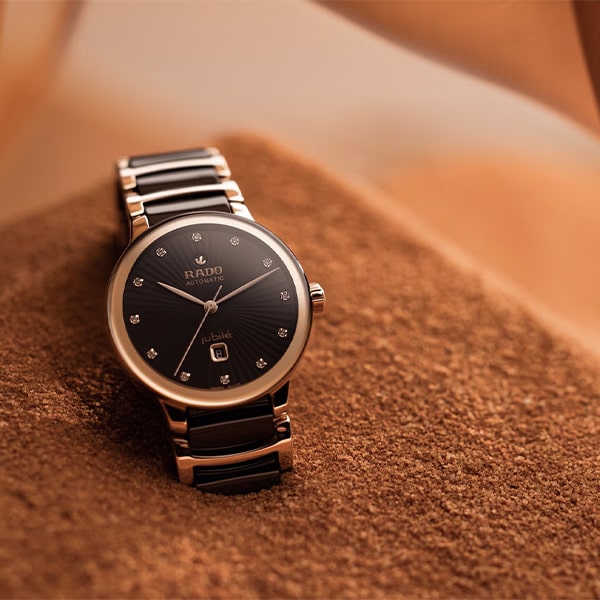 RADO © 2025 Eine Luxusuhr aus der Rado Centrix Kollektion mit schwarzem Zifferblatt und silbernen Akzenten auf einer strukturierten braunen Oberfläche. Die Uhr verfügt über ein silbernes Metallband und kleine, elegante Kristalle, die die Stunden markieren. Auf dem Zifferblatt sind „Rado“ und „Jubile“ zu sehen, was ihr anspruchsvolles Erscheinungsbild unterstreicht.